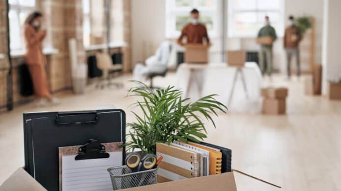 Focus on open cardboard box with office supplies, clipboards and plant against people moving into new office in background after a corporate relocation move